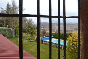 Vista a través de una ventana de metal que da a un patio trasero con piscina, terraza de madera y césped con árboles y arbustos.