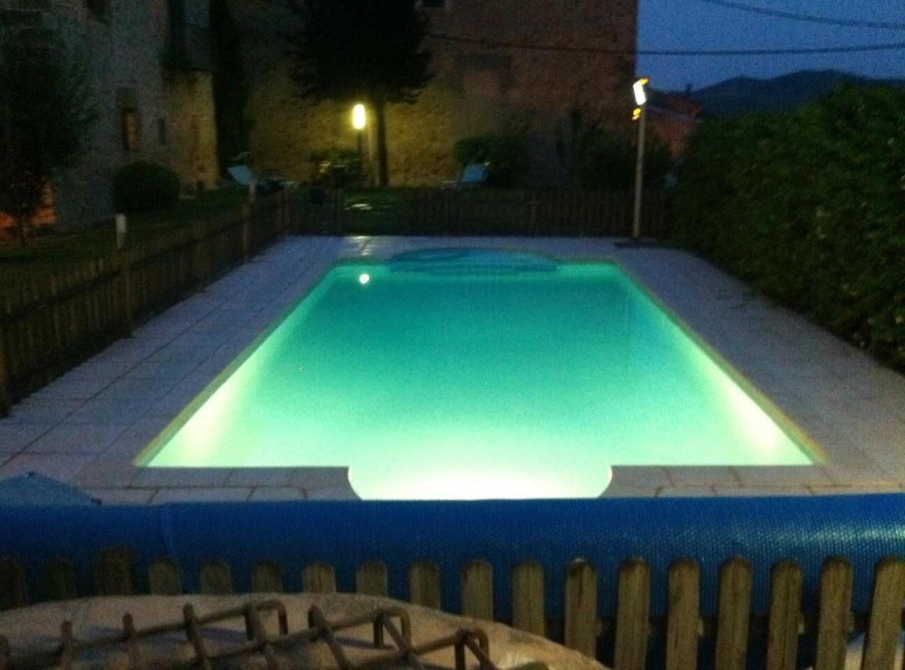 Una piscina rectangular con borde azul y parte interior azul claro, rodeada por una valla de madera y un suelo empedrado.