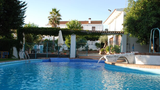 Une piscine avec un pont, entourée d’un bâtiment blanc doté d’un toit en tuiles et d’un patio couvert.