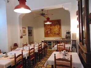 A dining area with multiple tables set for service, wooden chairs, a ceiling fan, and a large painting on the wall.