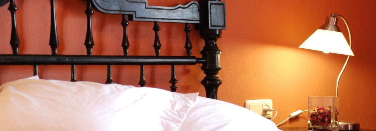 Una cama con ropa de cama blanca, cabecera de metal negro y mesita de noche con una lámpara y un recipiente de vidrio.