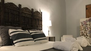 Un lit bien fait, agrémenté d’oreillers à rayures, d’une lampe de chevet et de serviettes pliées, le tout posé sur une couvre-lit de couleur foncée.