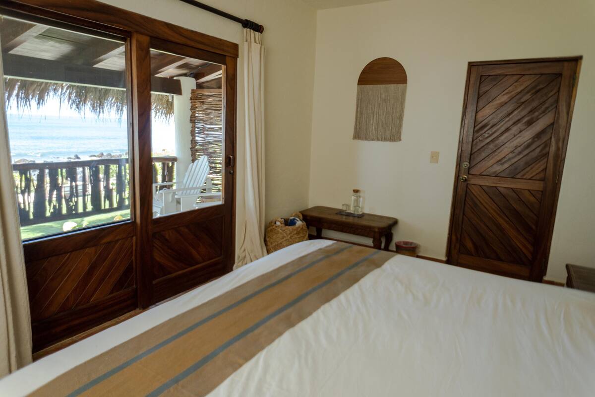 Une chambre à coucher avec un lit, une porte en bois, une petite table et une vue sur l’océan par une porte-fenêtre donnant sur un balcon.
