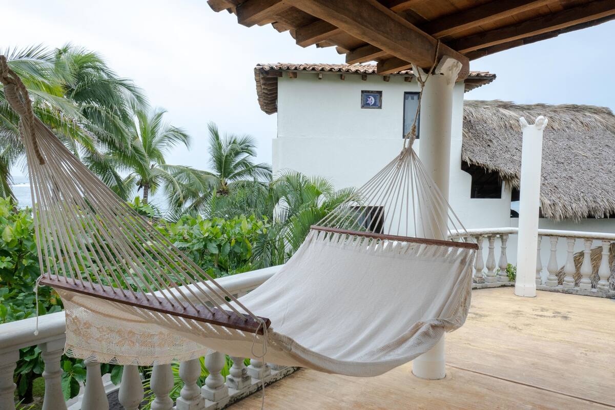 Un hamac sur un balcon, avec vue sur un bâtiment au toit de chaume et des palmiers.