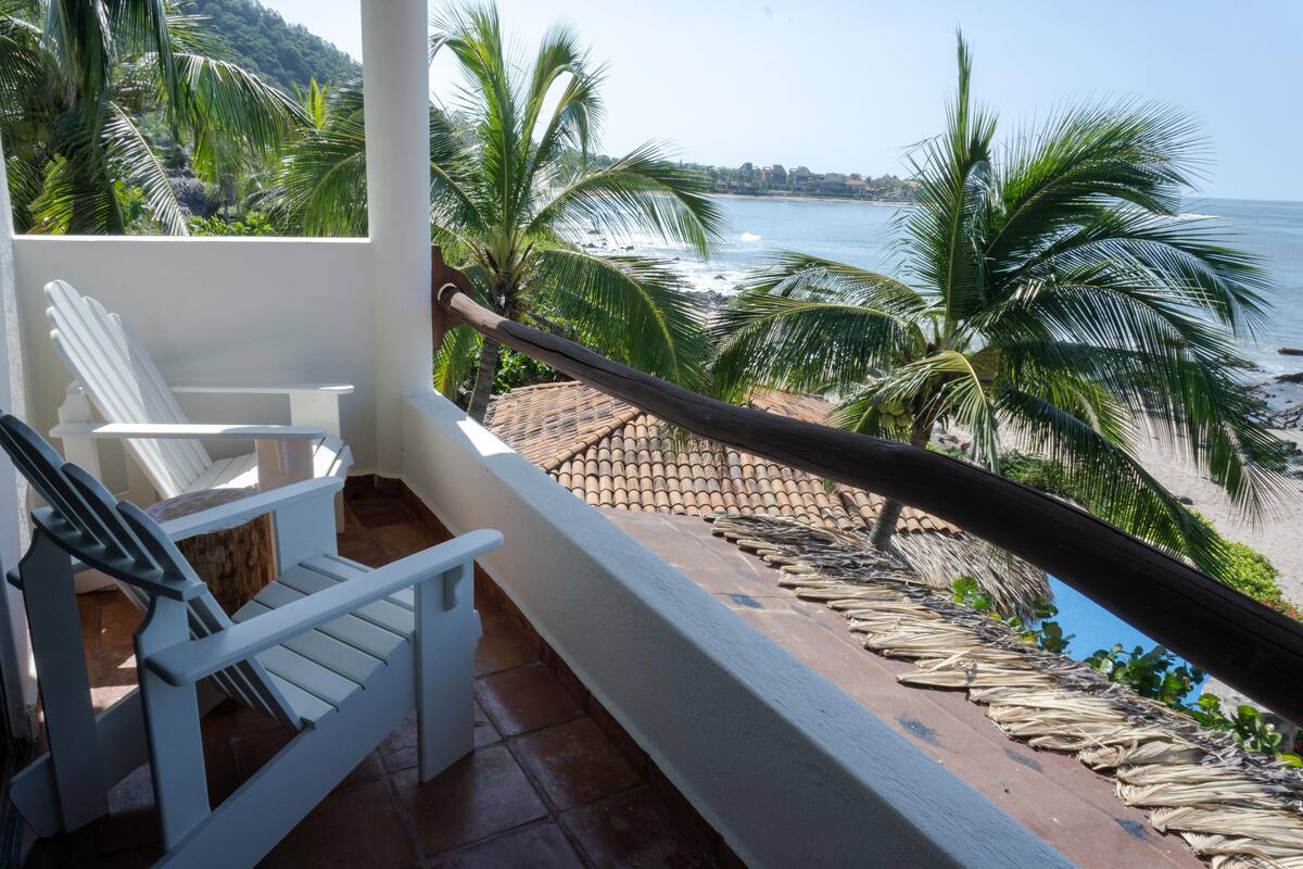 Un balcon avec des chaises blanches, une vue sur la plage, des palmiers et l’océan.