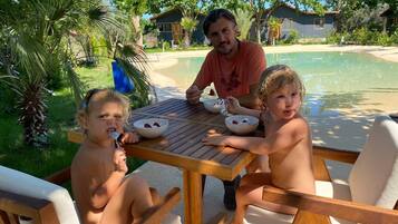 A family dining at a wooden table by a poolside, with a clear view of the pool and surrounding greenery.
