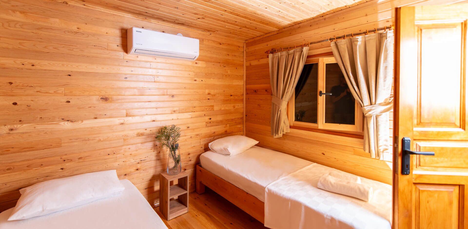 Una habitación de cabaña de madera con dos camas individuales, una mesita con un florero y una ventana con cortinas.