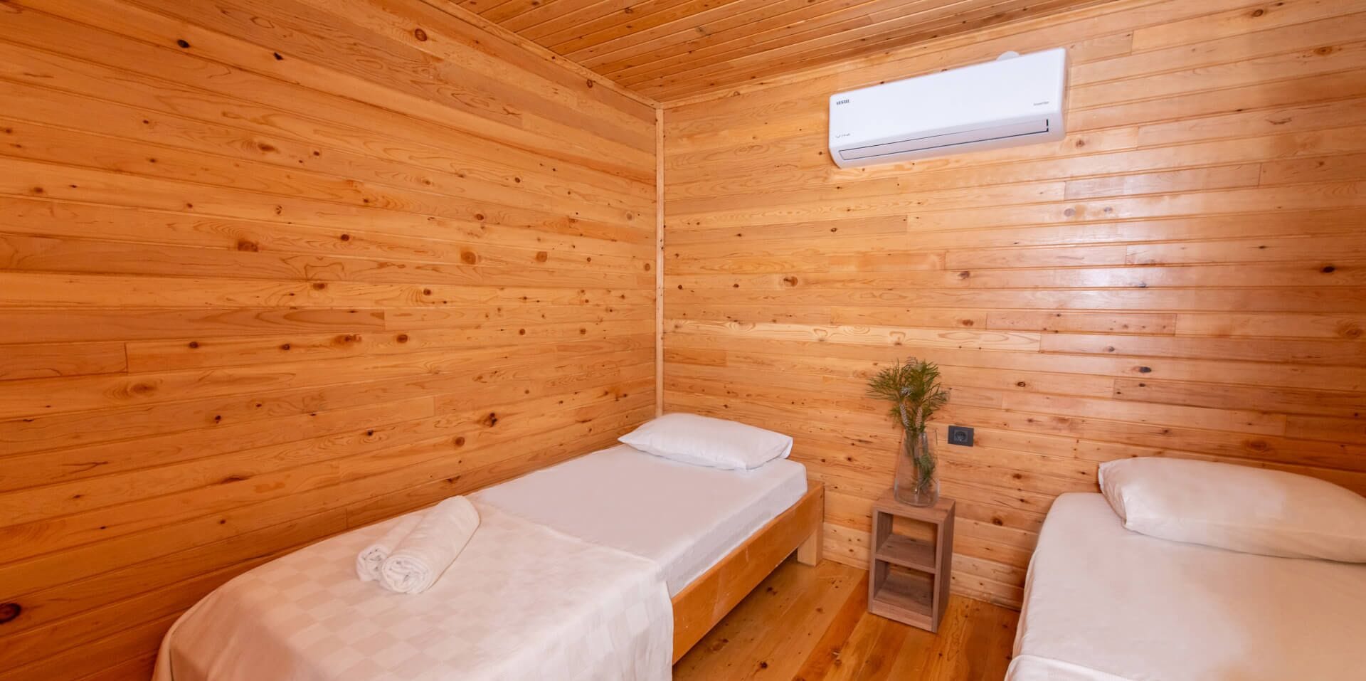 A small, wooden cabin room with two beds, a small table, and an air conditioning unit.