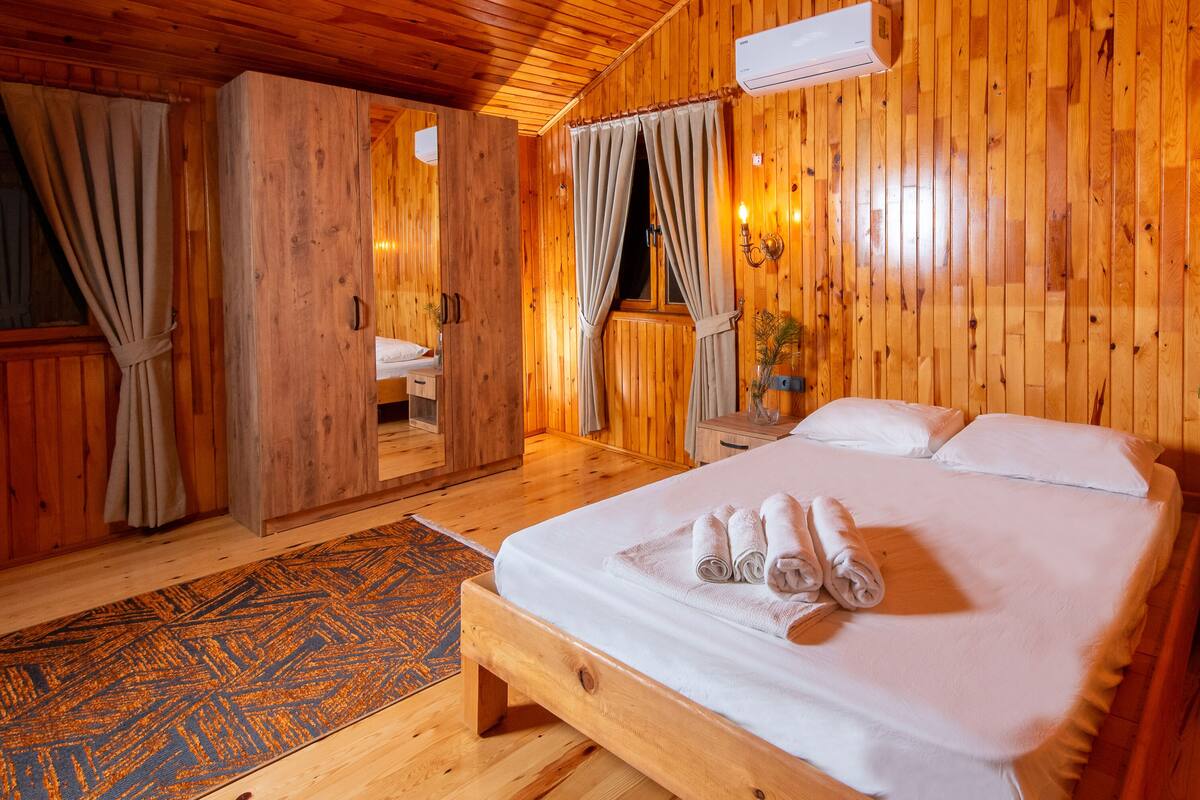 A wooden room with a bed, a nightstand, a mirror, and a window with curtains.