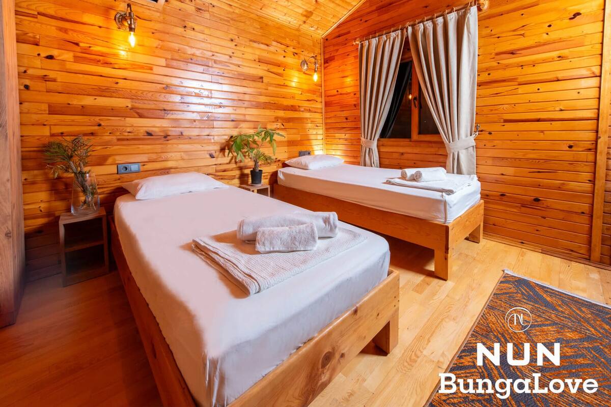 A wooden cabin bedroom with two beds, a potted plant, and a window with curtains.