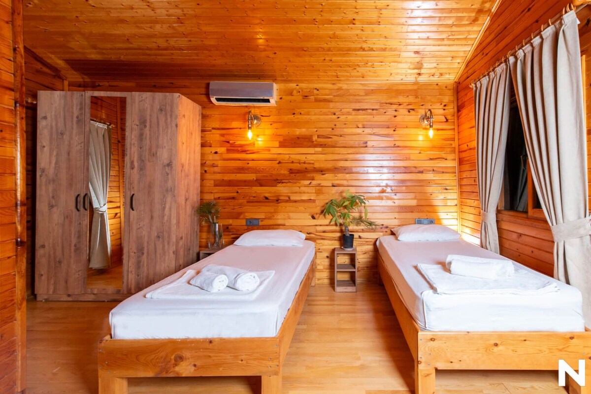 A wooden cabin room with two beds, a small table, and a window with curtains.