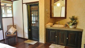 Quarto com cama, cadeira de madeira, porta de madeira escura, pia com espelho e uma pequena planta no balcão.