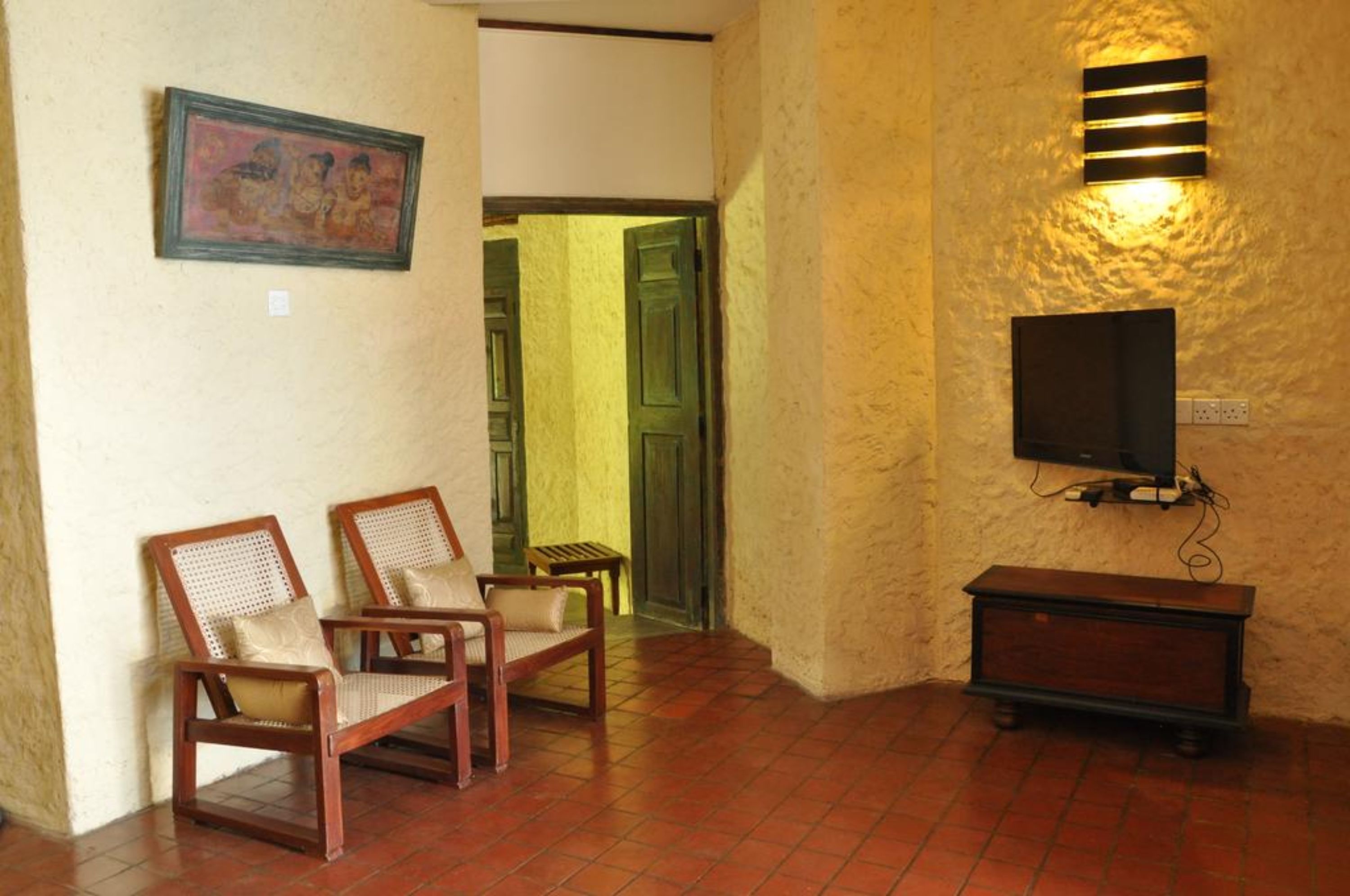 Una habitación con dos sillas de madera, una mesa pequeña, un televisor montado en la pared y una puerta que da a otra estancia.