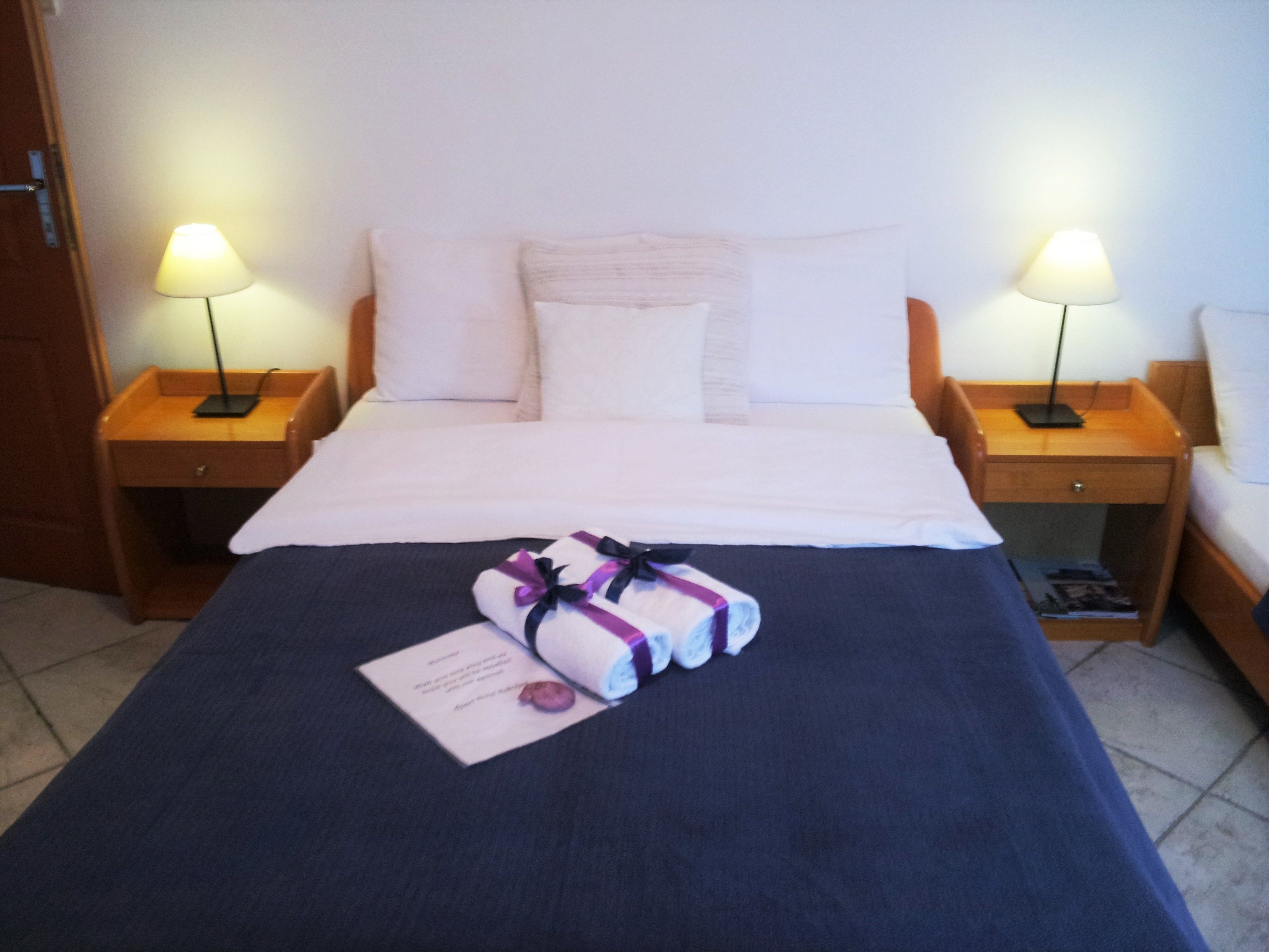 Una cama bien hecha con sábanas blancas, dos lámparas de noche y una toalla doblada con un lazo morado sobre la cama.