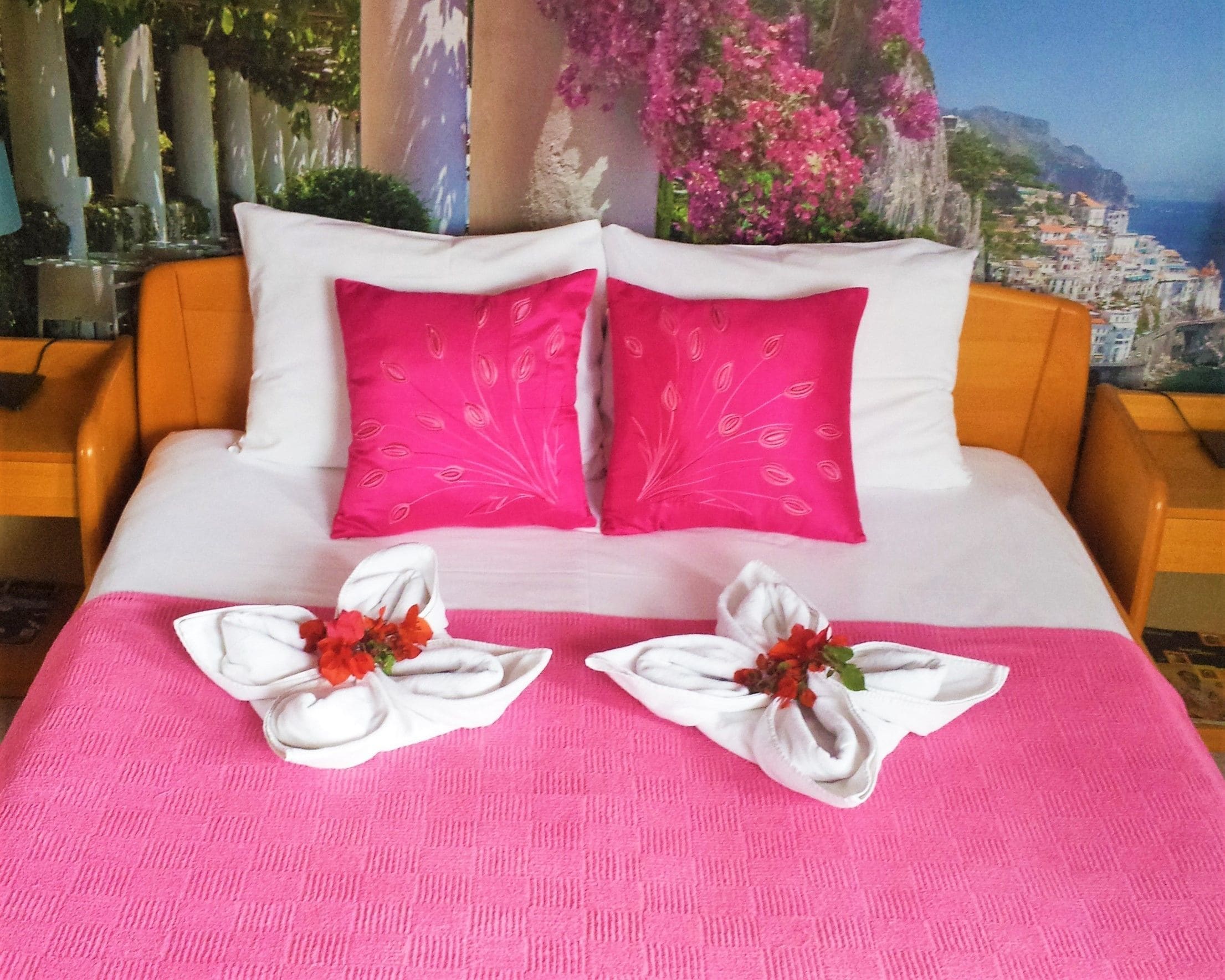 Een net opgemaakt bed met roze beddengoed en kussens, twee handdoeken gevouwen in bloemvormen, en een prachtig uitzicht op de kust op de achtergrond.