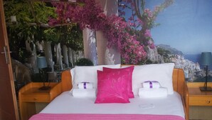 Un lit recouvert de draps blancs et d’une couverture rose, flanqué de tables de chevet en bois surmontées de lampes, le tout se détachant sur un mur orné d’une fresque représentant un paysage côtier avec des fleurs roses et un bâtiment.