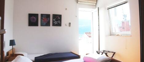 A small, bright room with two beds, a window with a view, and a wall-mounted air conditioner.