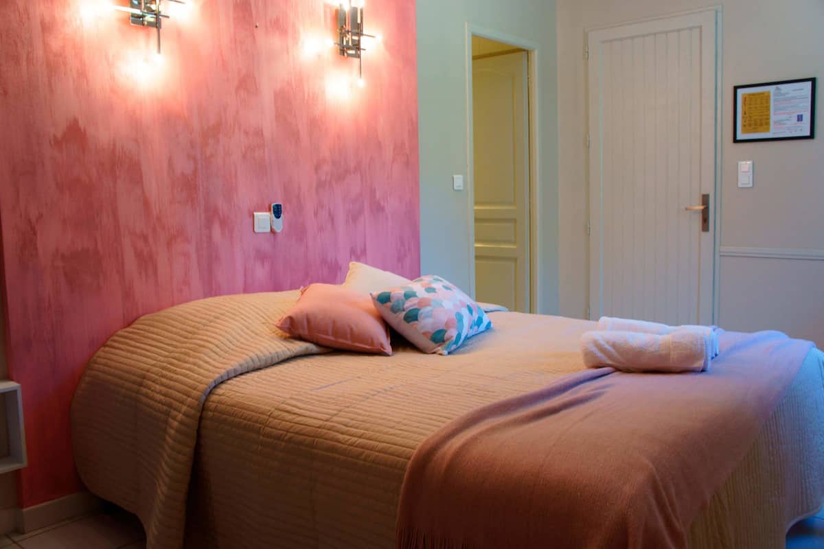 A bedroom with a pink wall, a bed with a quilted bedspread, two pillows, and a folded towel.