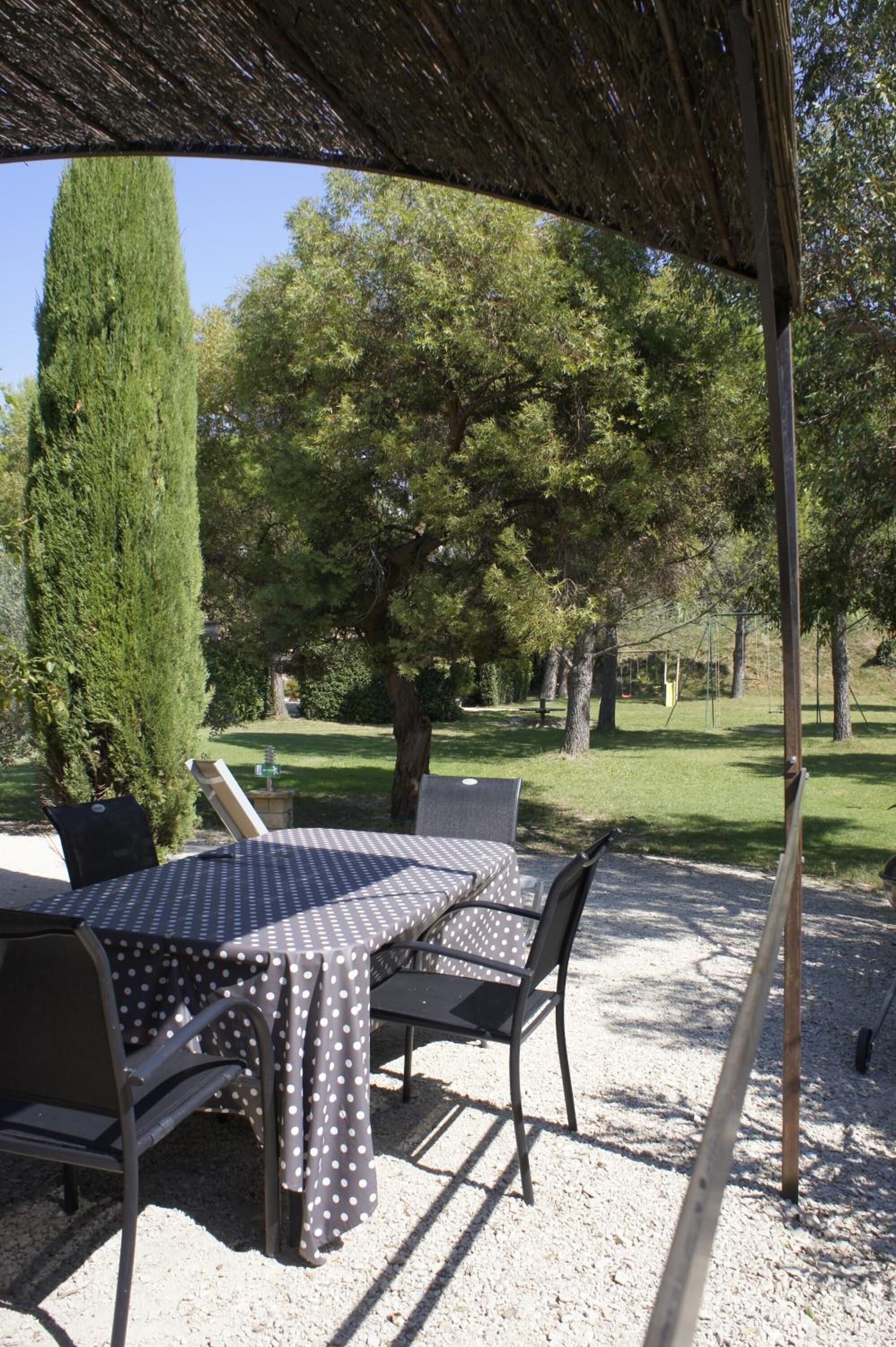 Un espacio de comedor al aire libre con una mesa y sillas bajo una pérgola, rodeado de árboles y un césped.