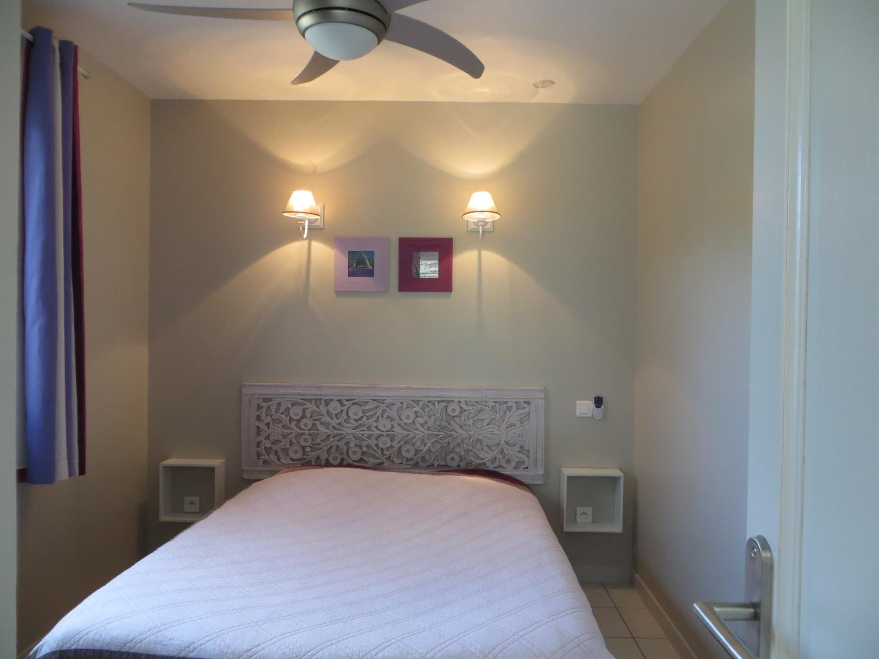 Une chambre d’hôtel comprenant un lit, un ventilateur de plafond, deux lampes fixées au mur, une tête de lit décorative et une petite table de chevet.