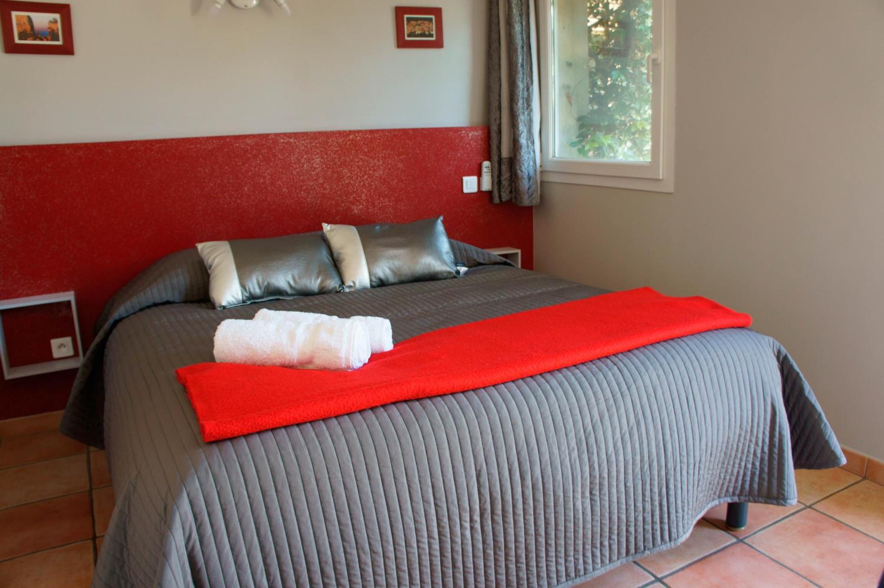Een netjes opgemaakt bed met een grijze deken, een rood hoofdbord en een rode plaid.