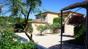 Une maison avec un toit en tuiles, entourée d’un jardin où poussent différentes plantes et agrémentée d’une pergola.