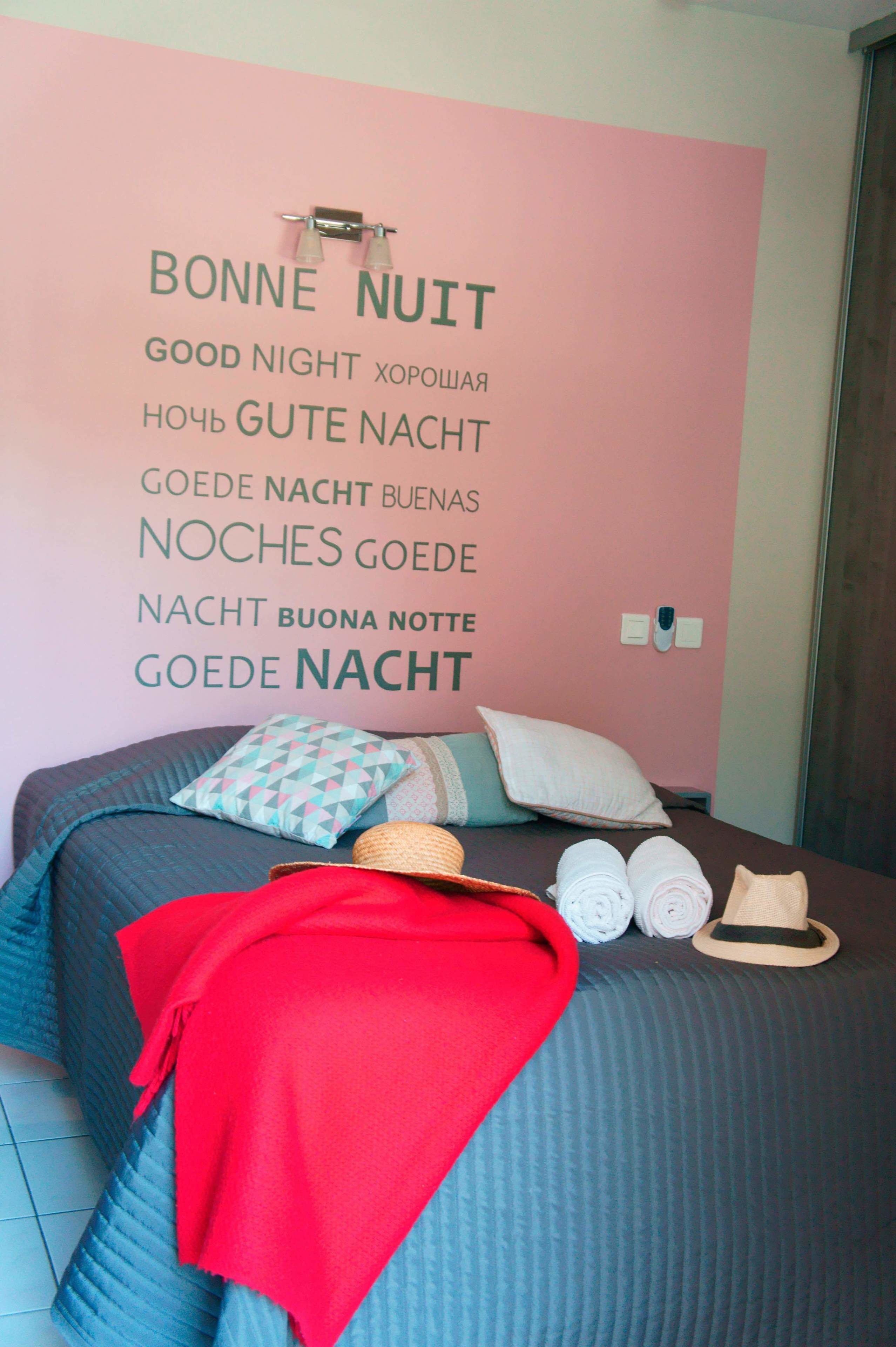 A bedroom with a pink wall featuring multilingual "Good Night" text. The bed has a teal comforter, multiple pillows, a red throw blanket, and rolled towels.
