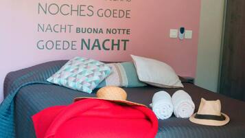 A bedroom with a pink wall featuring multilingual "Good Night" text. The bed has a teal comforter, multiple pillows, a red throw blanket, and rolled towels.