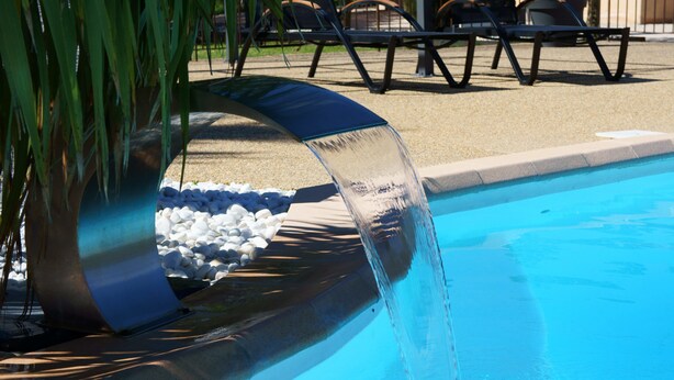 Une piscine agrémentée d’un élément décoratif aquatique, entourée de chaises longues et d’un palmier.