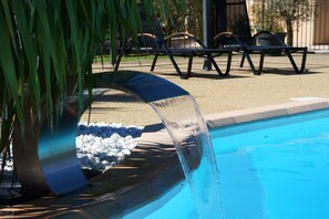 A pool with a water feature, surrounded by lounge chairs and a palm tree. - Hôtel Résidence Les Peupliers (Loriol-du-Comtat)