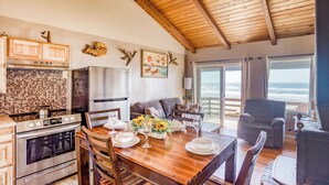 Une cuisine spacieuse avec un plafond en bois, un coin repas avec une table en bois dressée pour quatre personnes, et une vue sur l’océan grâce à de grandes fenêtres.