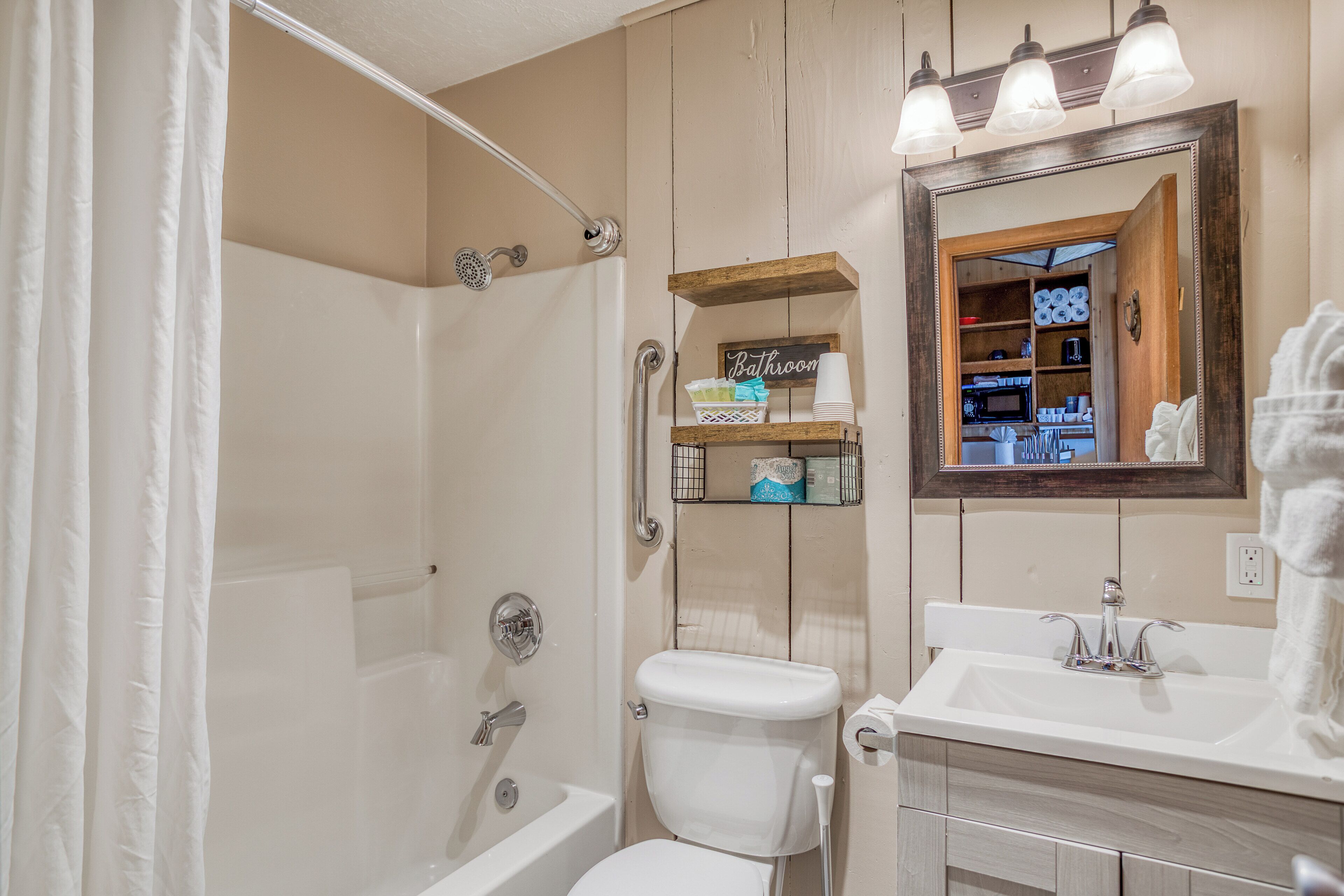 A bathroom with a shower, toilet, and sink. There is a mirror above the sink and a shelf with toiletries.