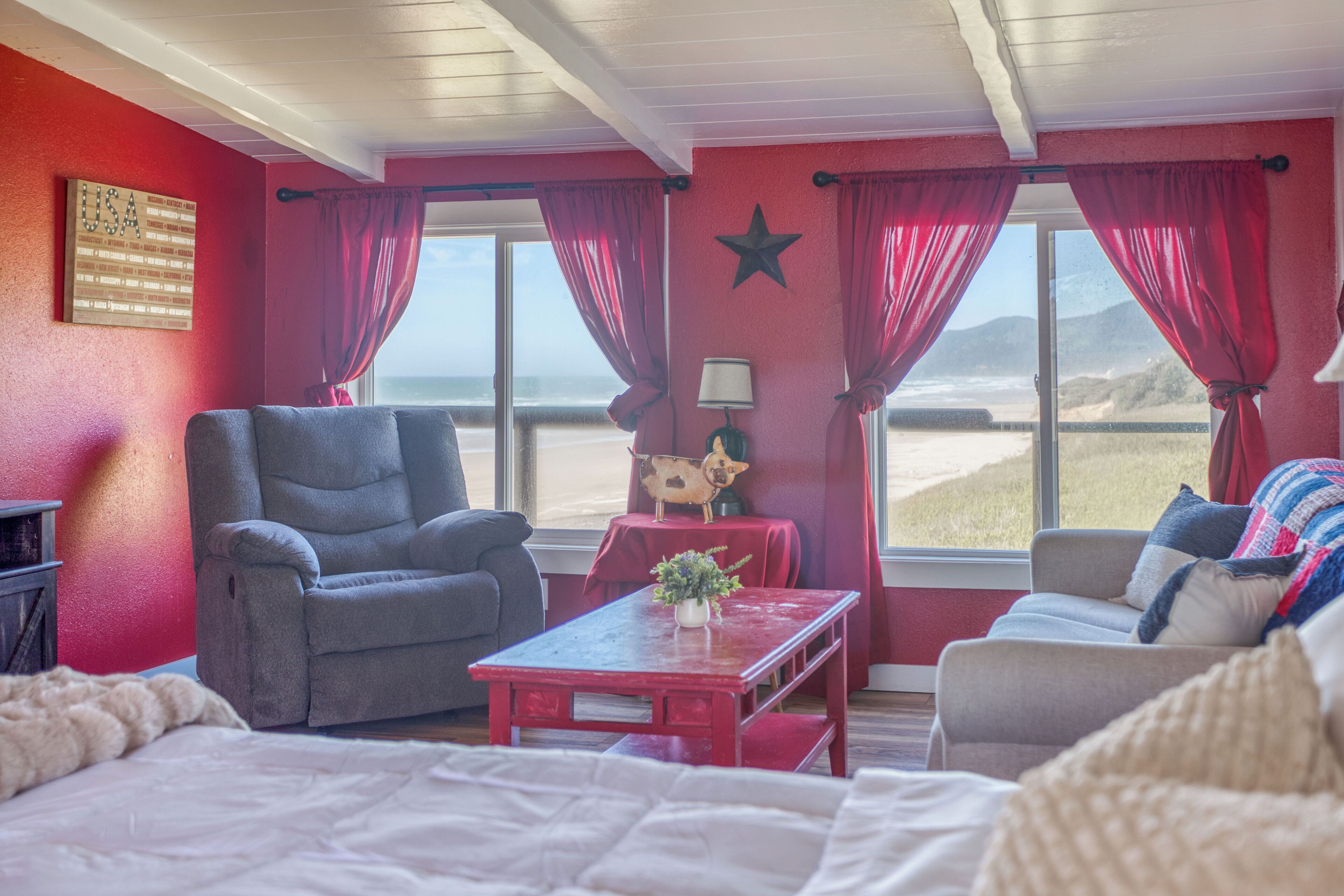 Une pièce comprenant un lit, un fauteuil gris, une table rouge avec une lampe, une petite figurine de chien et une vue sur la plage, visible à travers de grandes fenêtres ornées de rideaux rouges.