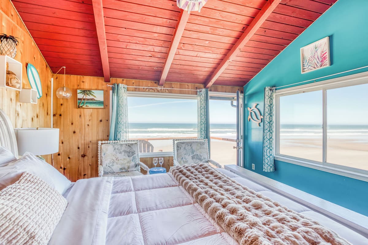 A room with a bed, a window with a beach view, a wooden ceiling, and a wall with a painting.