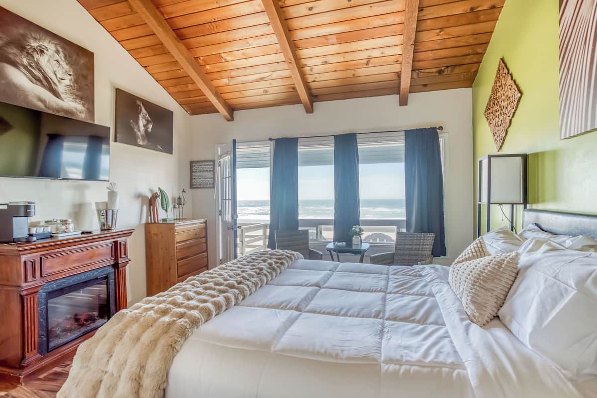 A bedroom with a large bed, a fireplace, a balcony with a table and chairs, and a view of the ocean.