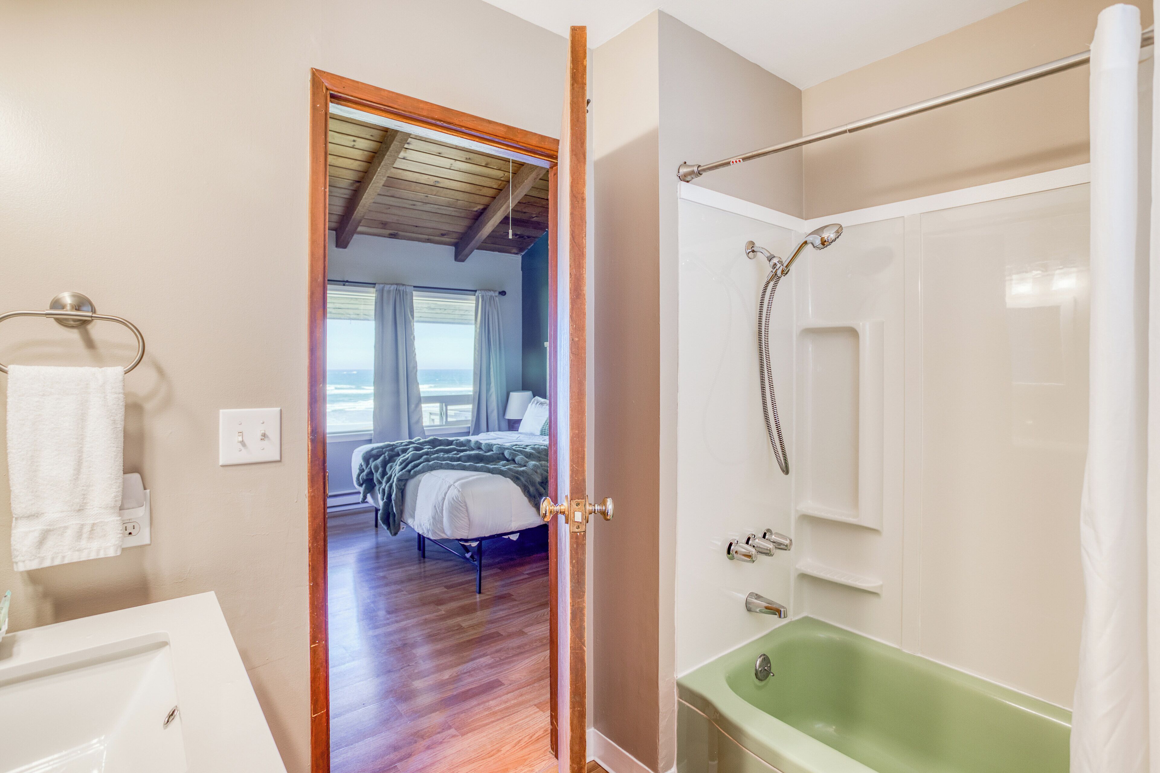A bathroom with a green bathtub, a shower, and a visible bedroom with a bed and window.