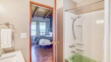 A bathroom with a green bathtub, a shower, and a visible bedroom with a bed and window.