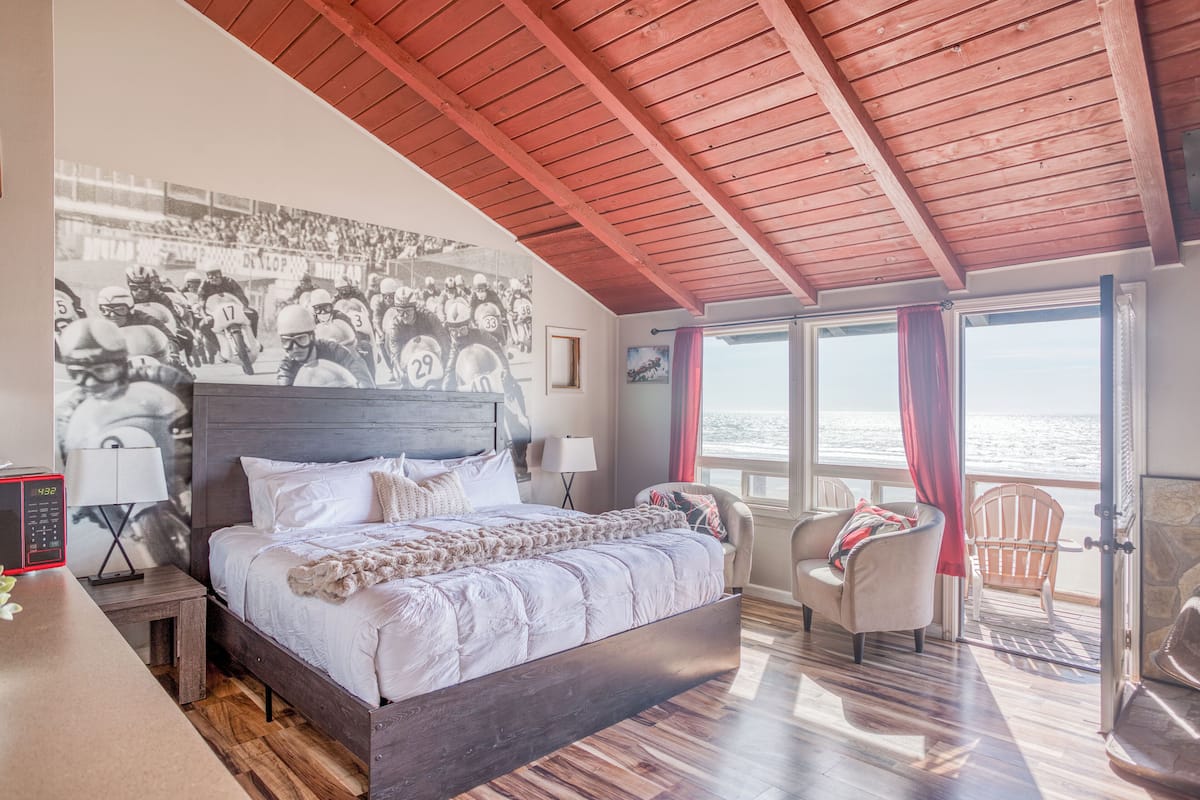 A bedroom with a large bed, a wooden ceiling, a view of the sea, a chair, and a nightstand with a lamp.