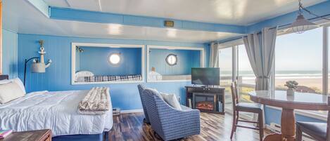 Una habitación con cama, sofá, televisor y una mesa junto a un ventanal con vistas a la playa.