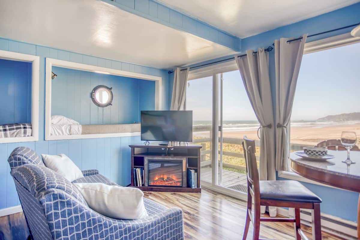 A room with a blue wall, a television, a fireplace, a sofa, a dining table, and a view of the beach.