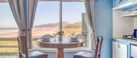 A dining area with a table and chairs, a window with curtains, and a view of the beach.