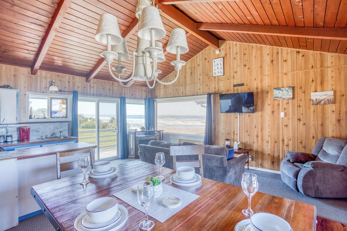 A wooden interior with a dining area, kitchen, and seating area, featuring large windows and a television.
