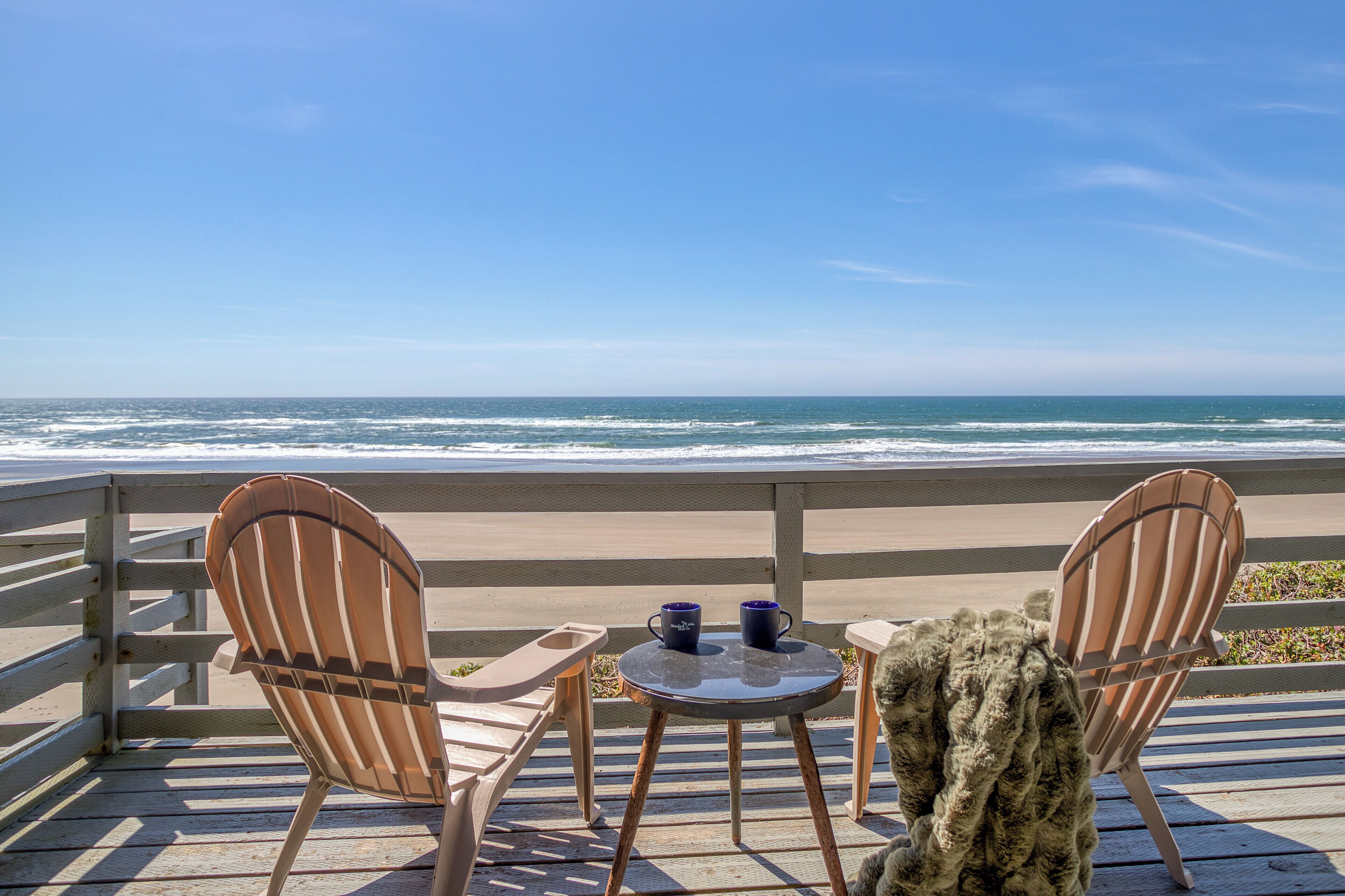 Una terraza de madera con dos sillas Adirondack y una mesita redonda, con vista a una playa y el océano al fondo.