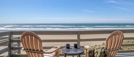 A wooden deck with two Adirondack chairs and a small round table, overlooking a beach with the ocean in the background.