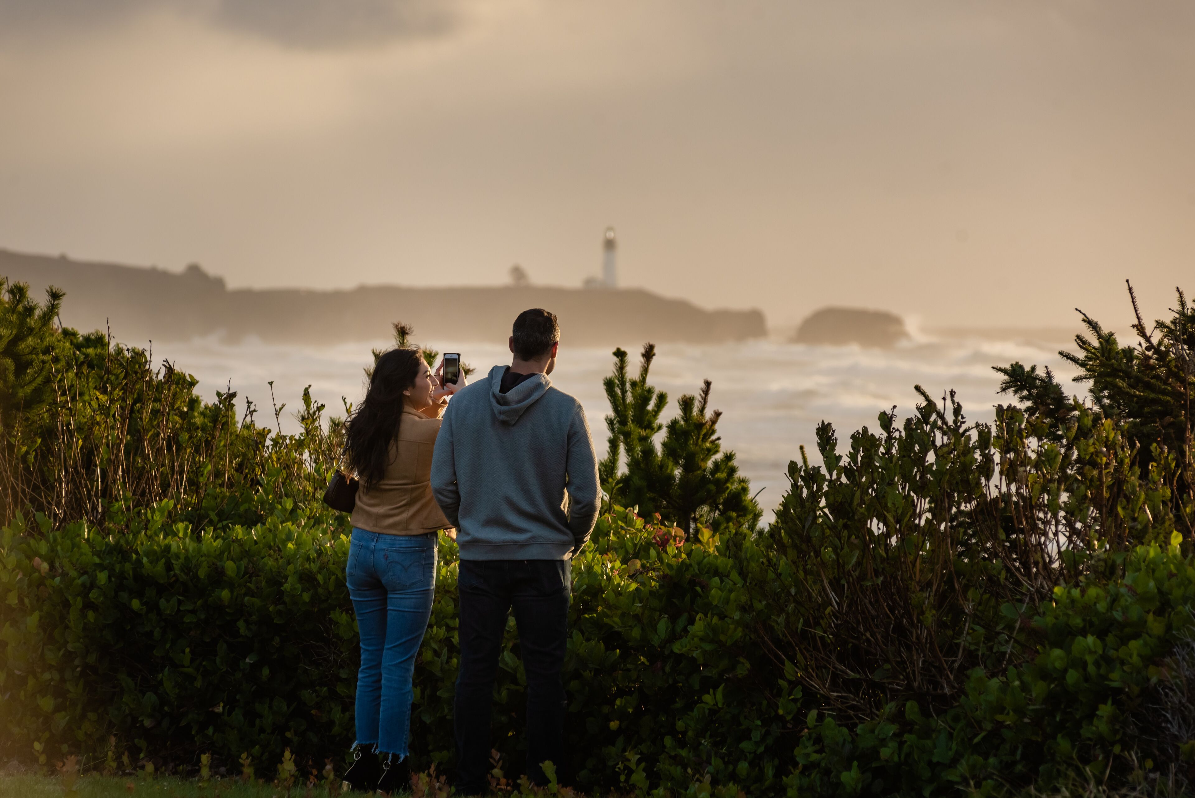 Two people standing by a coastal area with a lighthouse in the background.