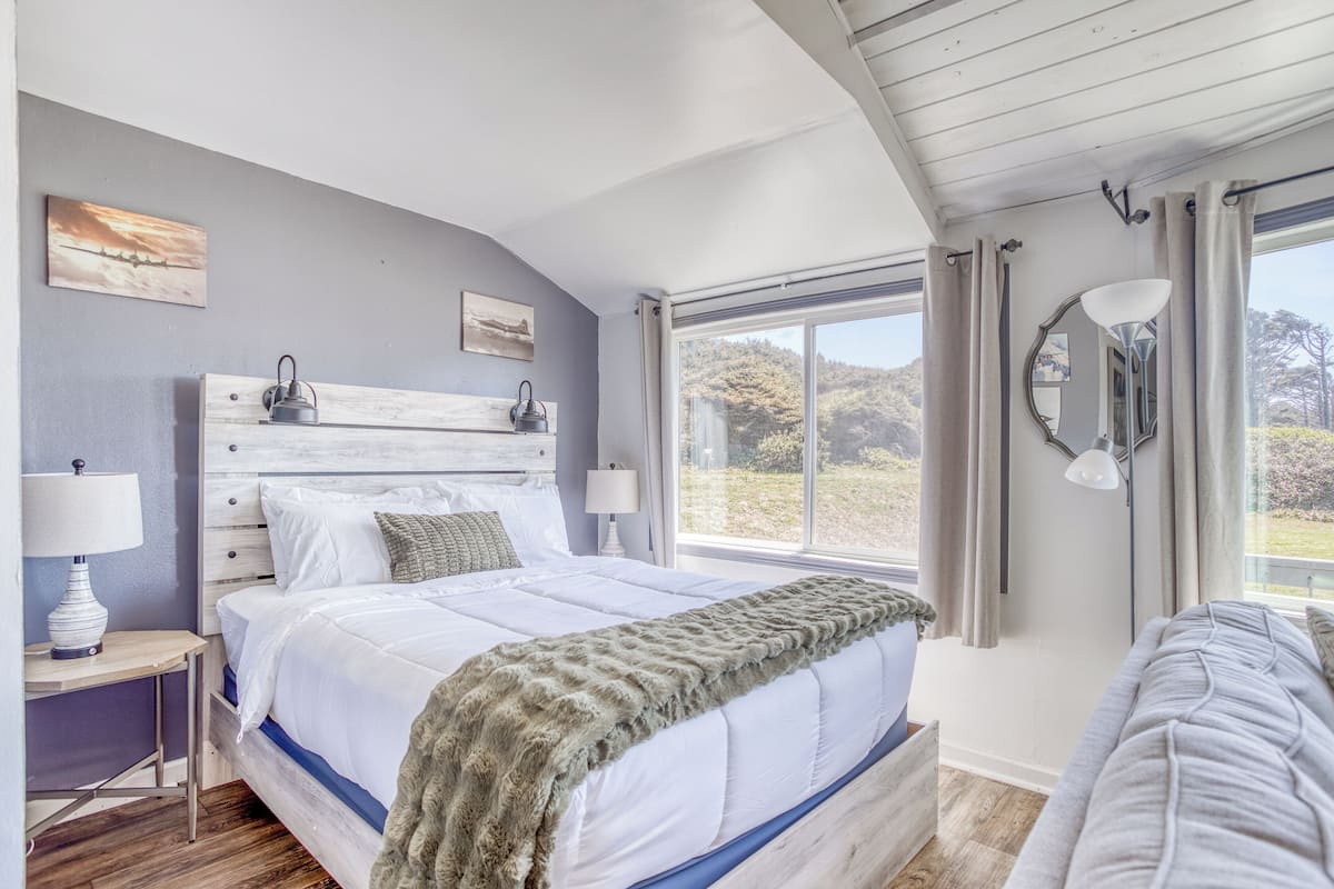 A bedroom with a large bed, a bedside table with a lamp, a window with curtains, and artwork on the wall.