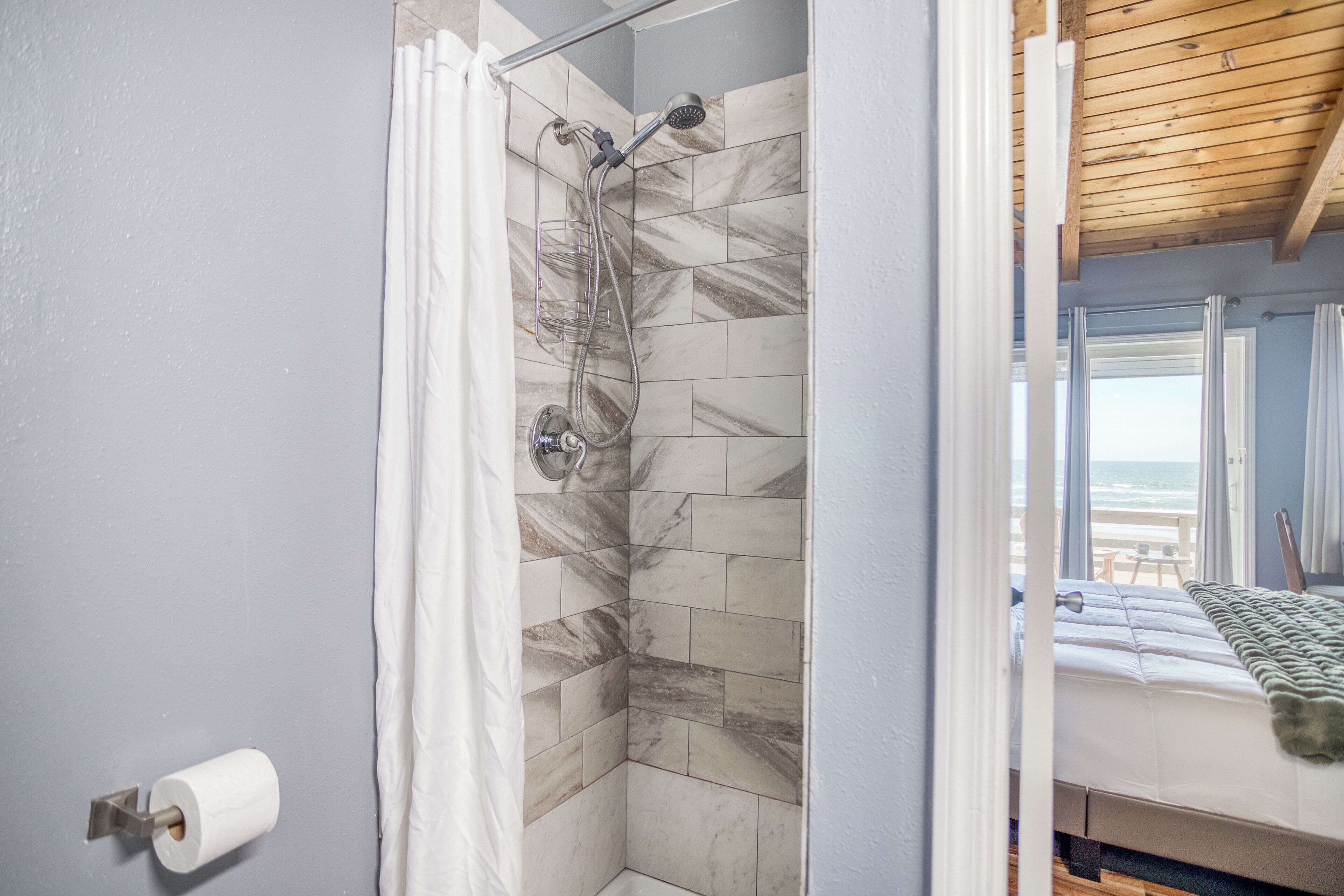 A bathroom with a shower, marble tiles, a toilet paper holder, and a bed with a view of the ocean.