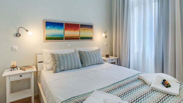 A neatly made bed with striped pillows, a bedside table with a book and a bottle, and a wall with three framed paintings. - Casa 1926 (Chania)