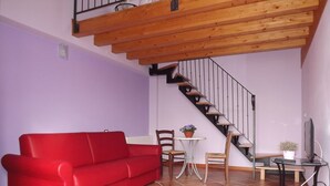 A living room with a red sofa, a small round table, two chairs, a potted plant, a TV, and a wooden staircase. - Agriturismo Cascina Rossano (Provaglio d'Iseo)