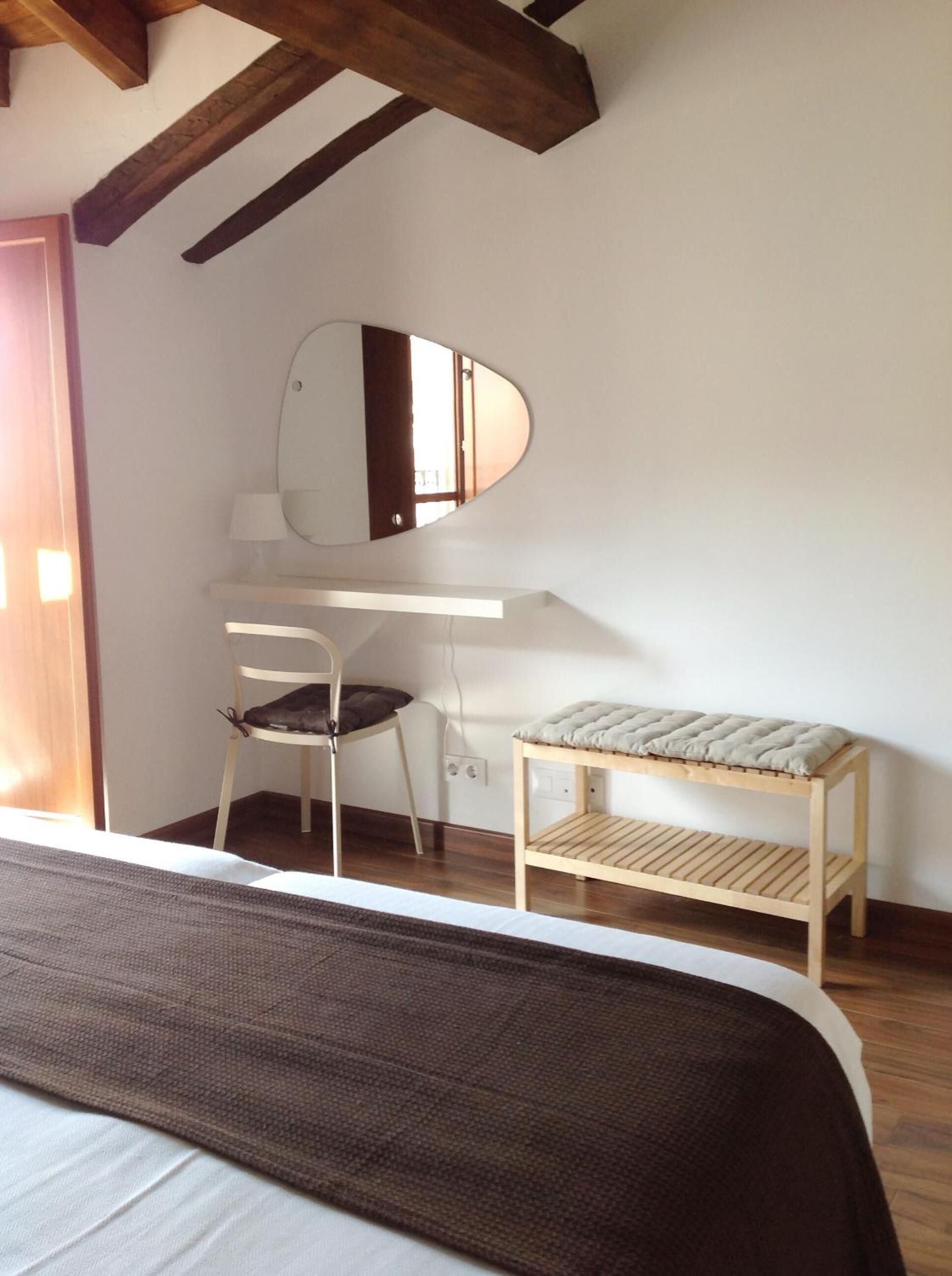 Un dormitorio con una cama, un escritorio, una silla, un taburete pequeño y un espejo.